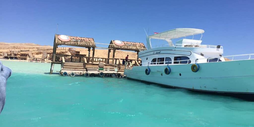 A boat docked beside a wooden pier at Orange Bay Marina, Hurghada, surrounded by calm turquoise waters and serene island vibes.