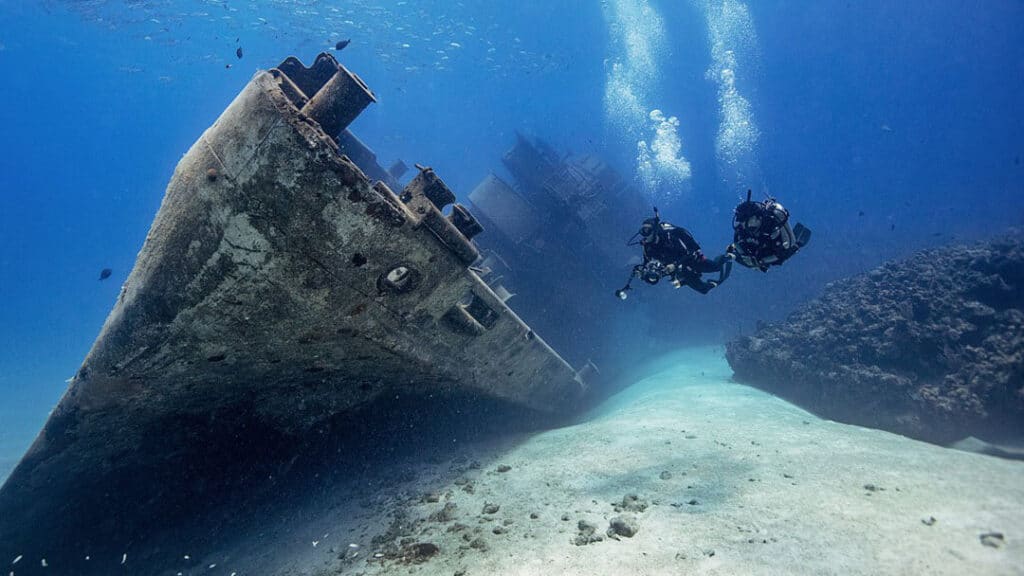 Two scuba divers explore the depths near a sunken ship, surrounded by marine life and underwater scenery.