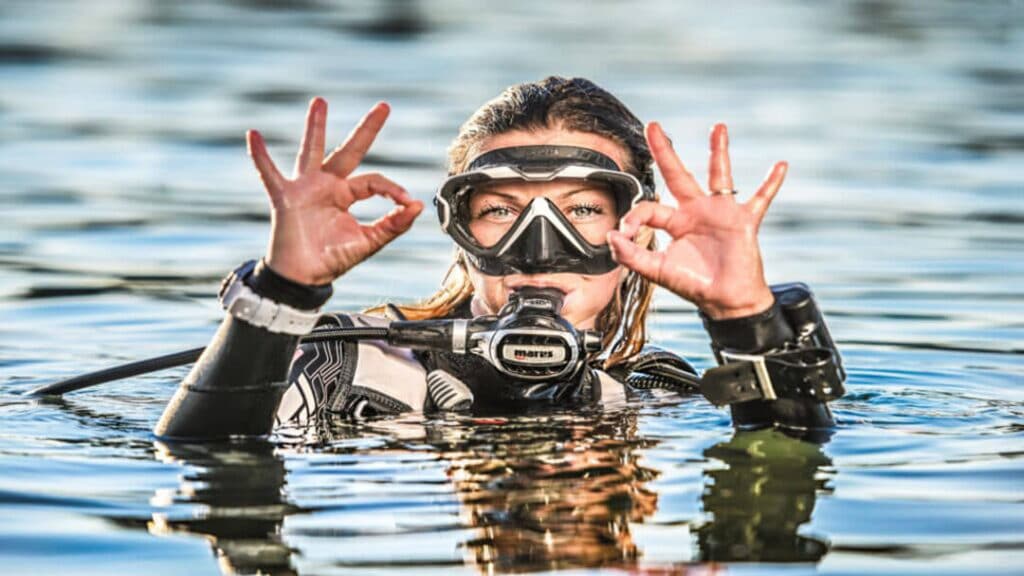 A woman wearing a scuba suit and mask is making the "OK" sign, indicating her comfort and safety while diving.